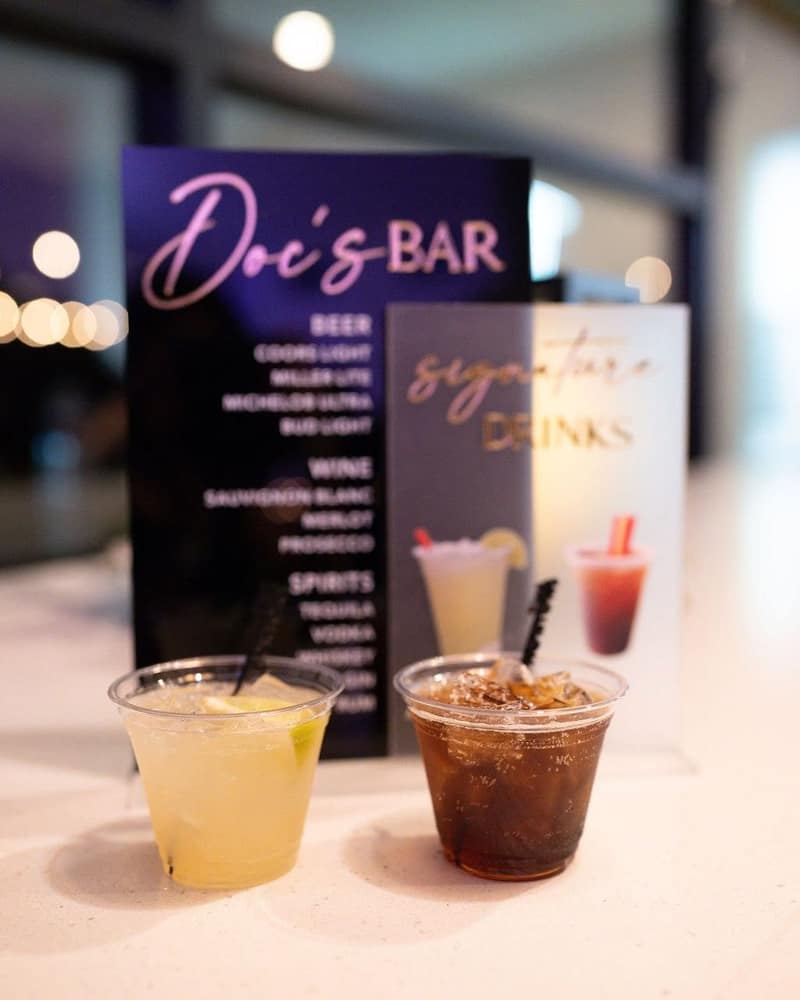 Illuminated bar menu listing the beer, wine and spirits on offer, with two cocktails poured in front of it