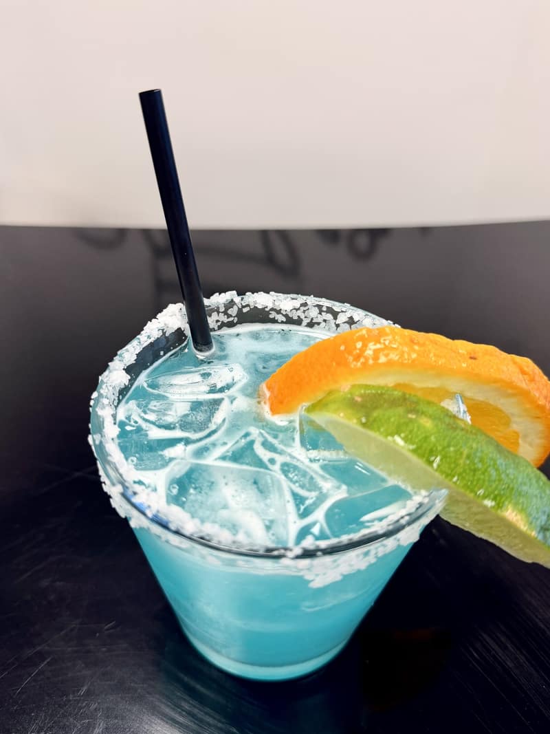Blue cocktail with a salted rim garnished with an orange slice and a lime wheel
