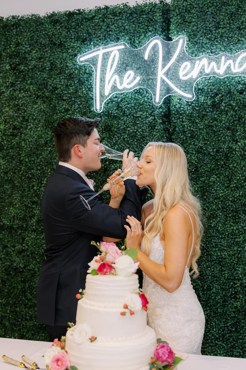 Couple toasting in front of a neon name sign and their wedding cake