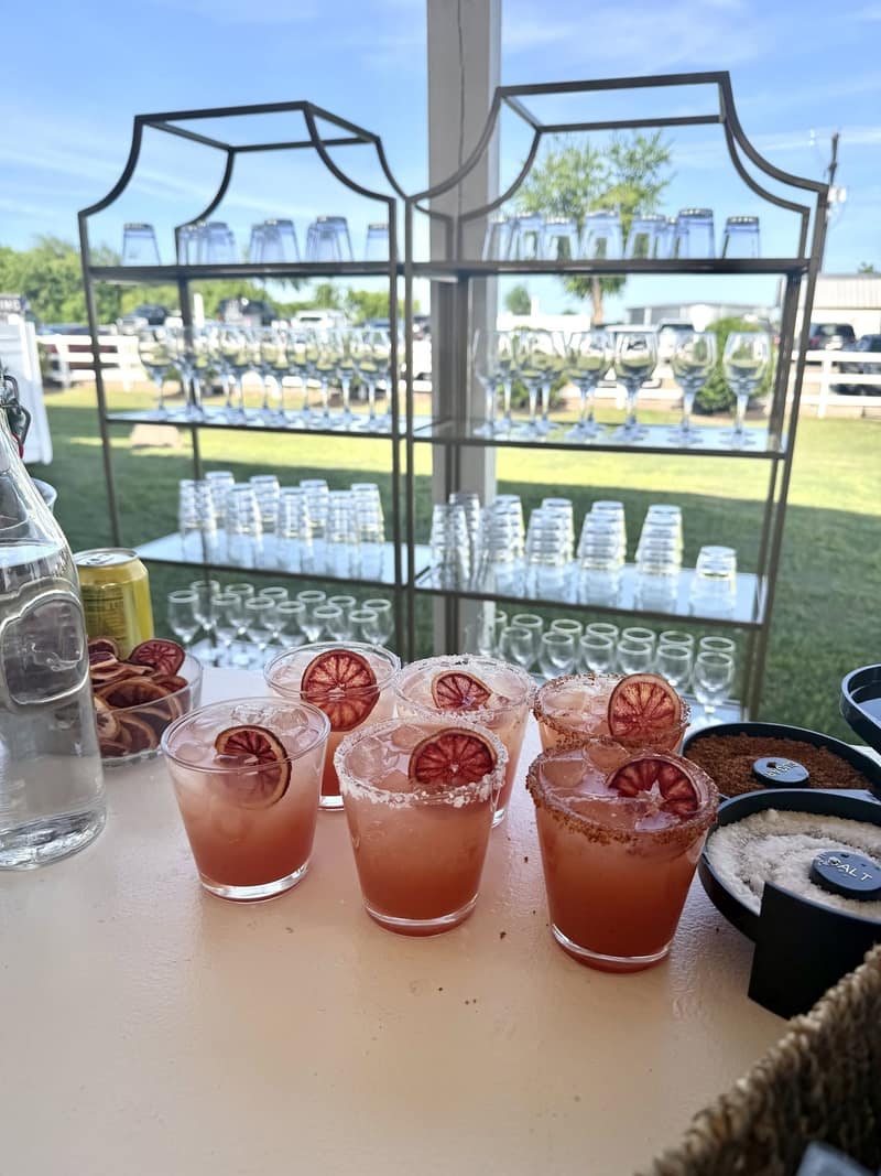 Racks of glassware on a metal shelf at an outdoor bar, with margaritas garnished with dehydrated blood orange lined up in front