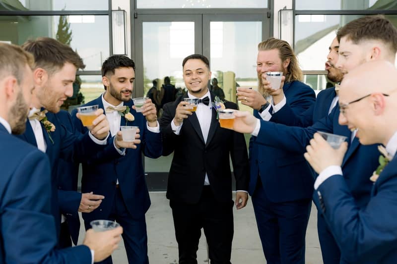 Groomsmen in blue suits crowding in to cheers the groom with cups of beer