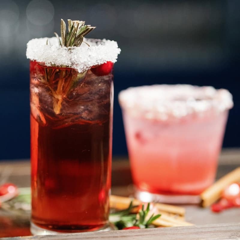 Two holiday cocktails with sugared rims, rosemary and cranberry garnishes on a wooden bar