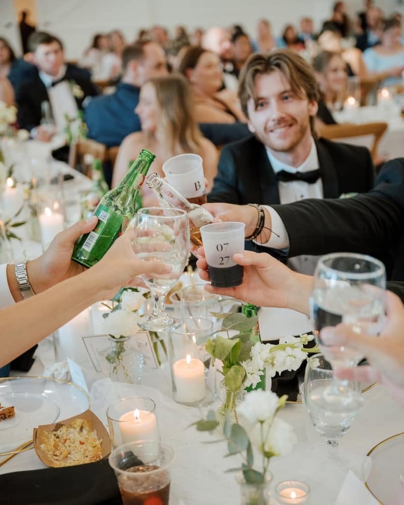 Wedding guests clinking cups at a candlelit reception table while a bottle is poured